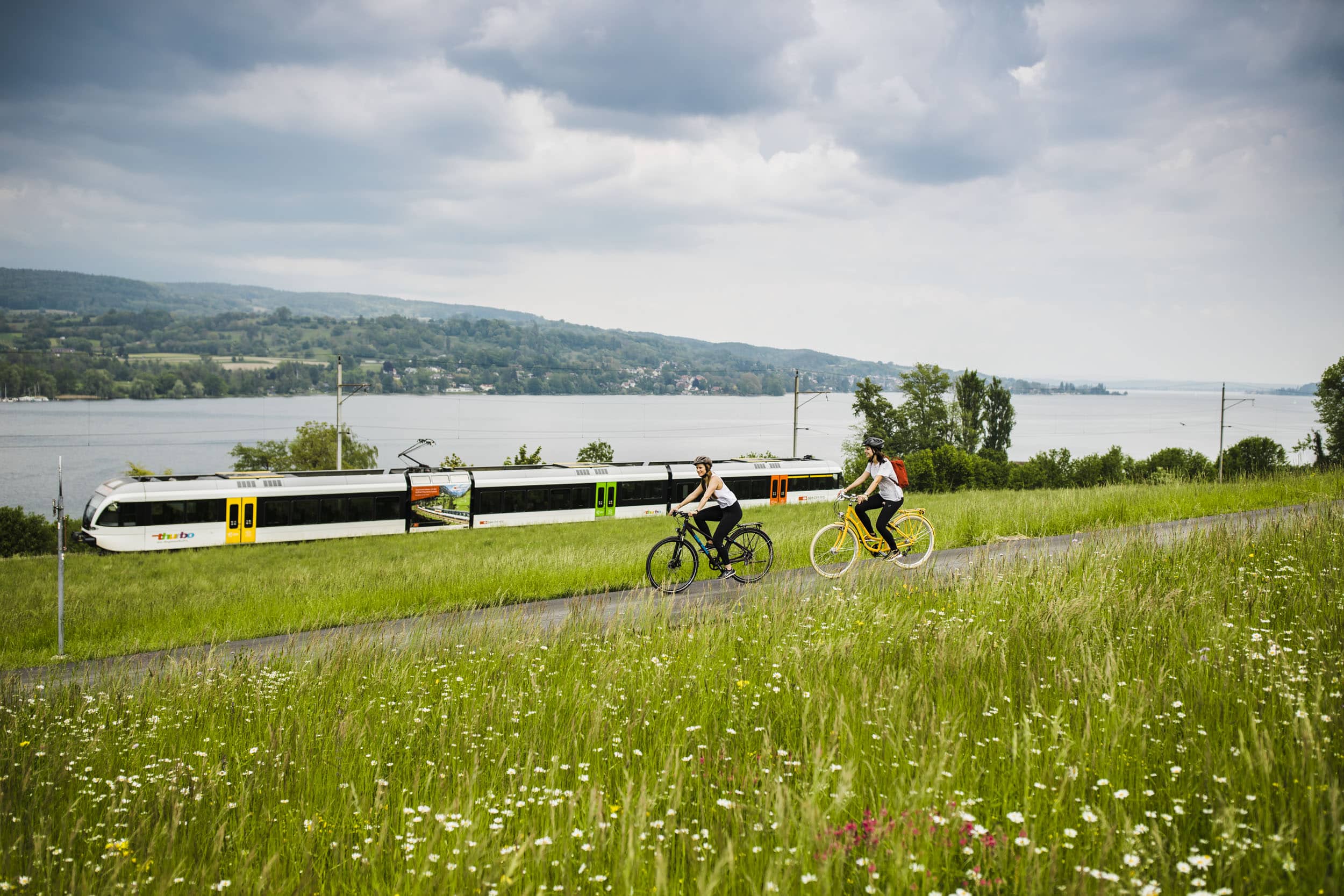 Zwei Radfahrer fahren auf einem Weg neben einer Bahnstrecke am See