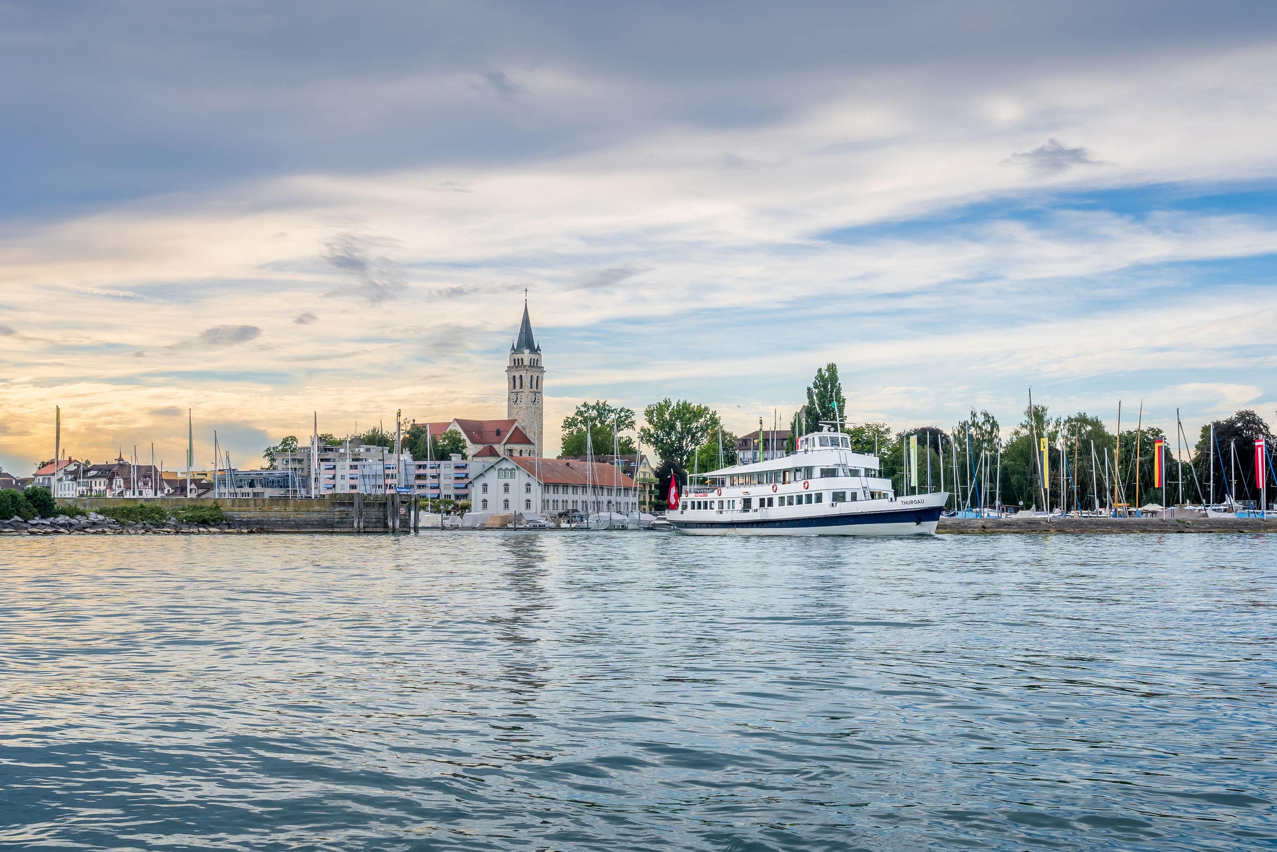 Fähre auf ruhigem See vor einer Stadt mit Kirche und Segelbooten