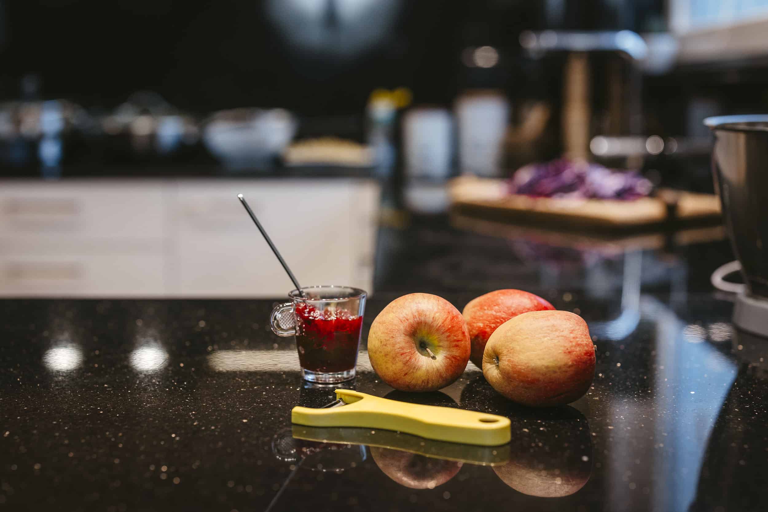 Fresh apples, yellow peeler, and glass of red jam on black kitchen counter