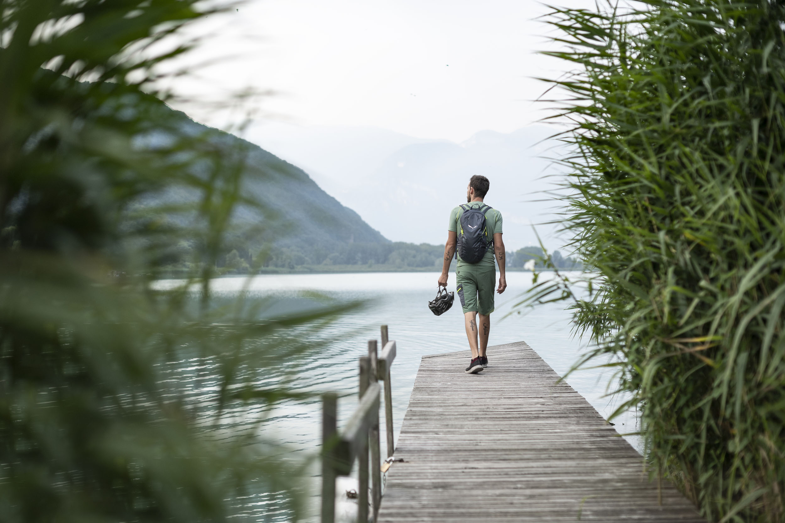 Mann mit Rucksack und Fahrradhelm geht auf Steg am See mit Bergen im Hintergrund