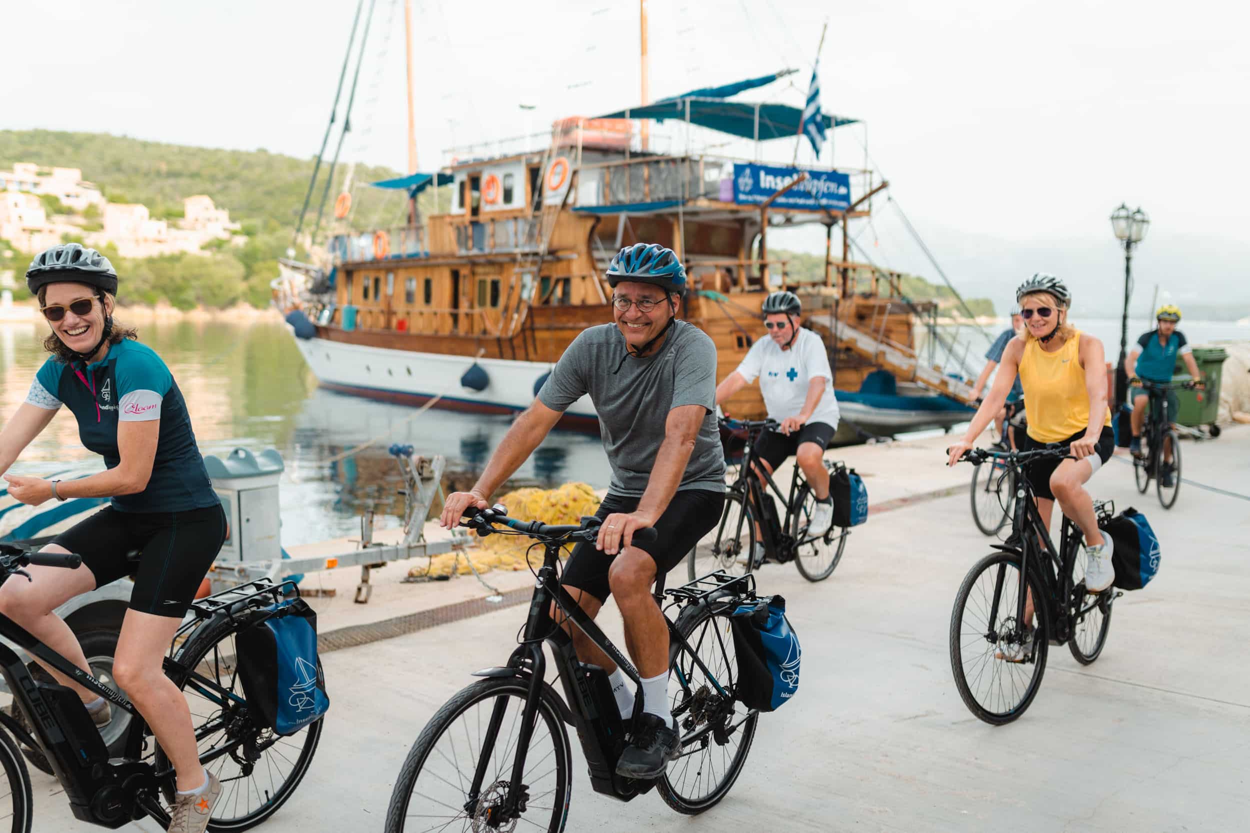 Gruppe von Radfahrern mit Helmen entlang eines Kai mit einem Holzboot im Hintergrund