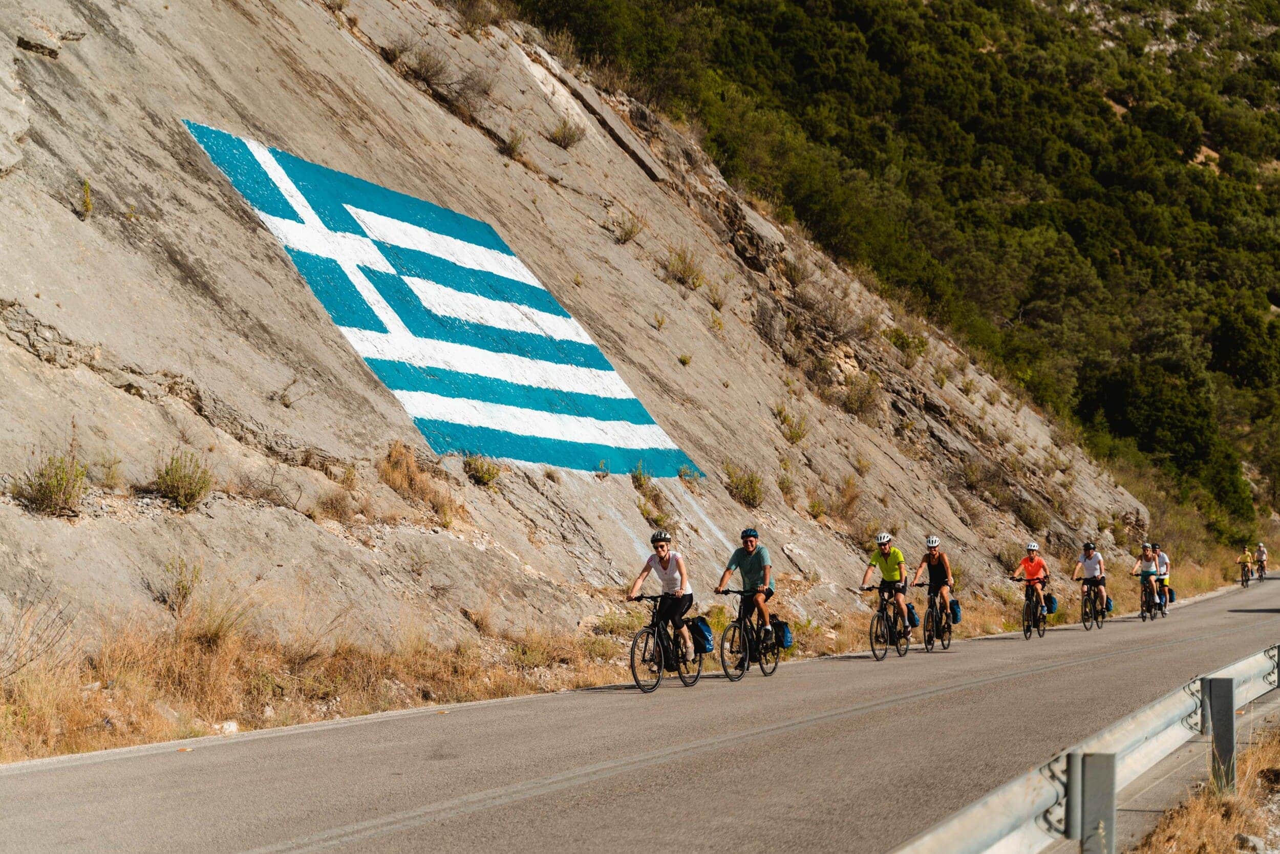 Fahrradfahrer auf Straße neben Felswand mit griechischer Flagge