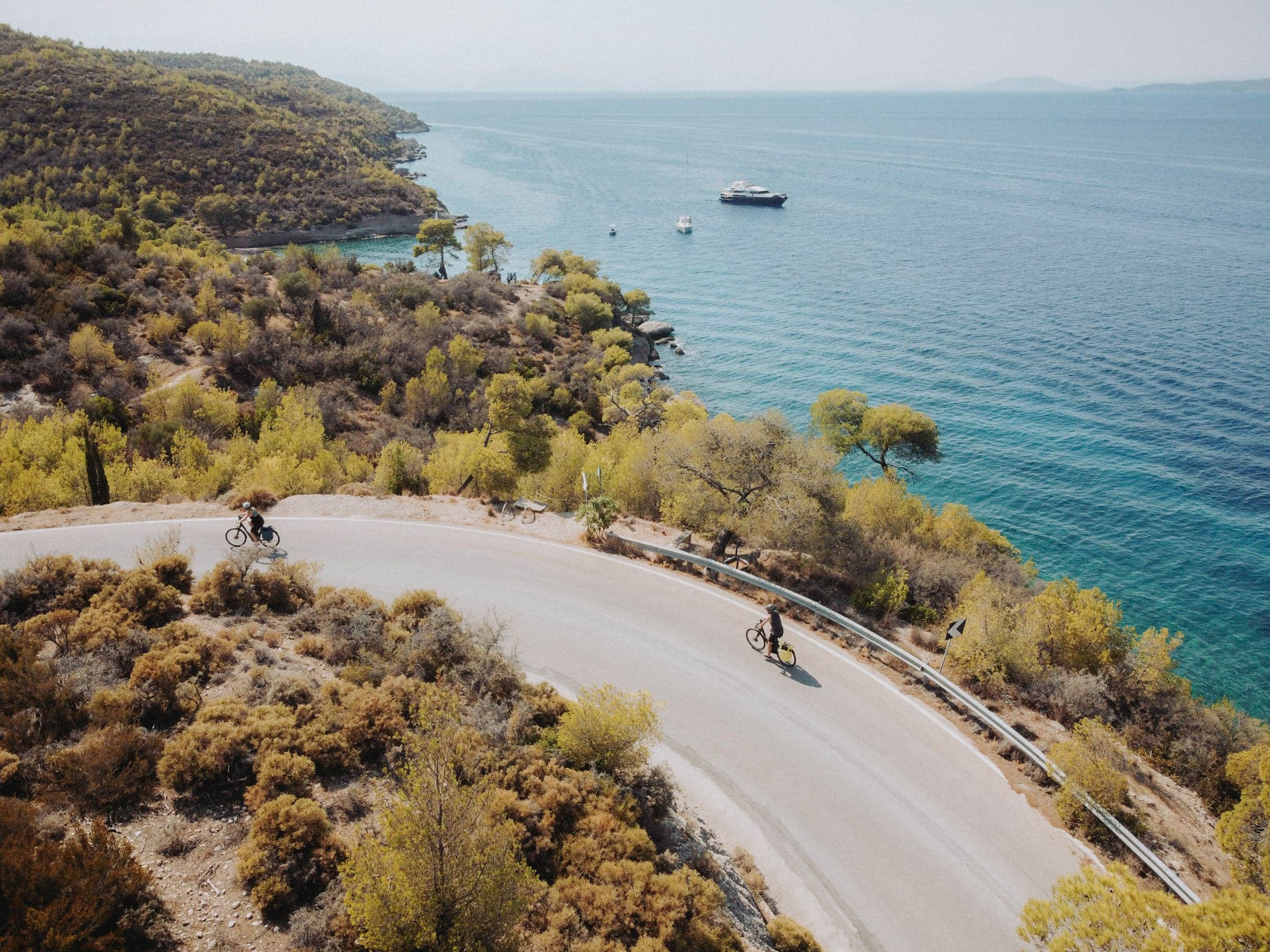 Radfahrer auf kurviger Straße am Meer mit bewaldeten Hügeln im Hintergrund